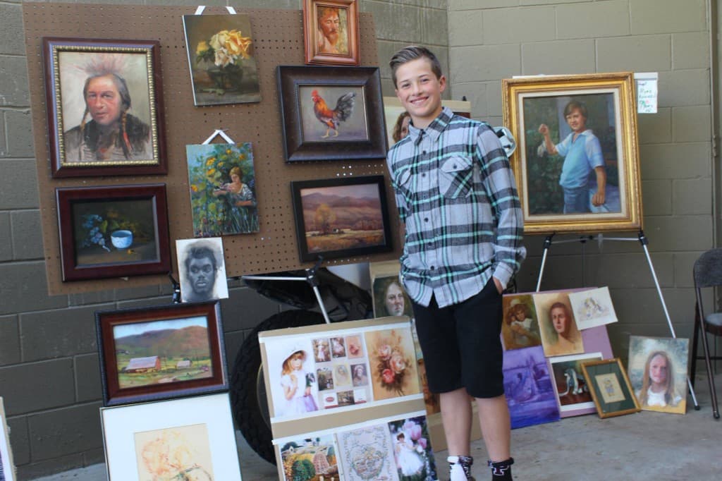 A young boy smiling in front of a large display of framed portraits, landscapes, and still lifes on pegboard and easels