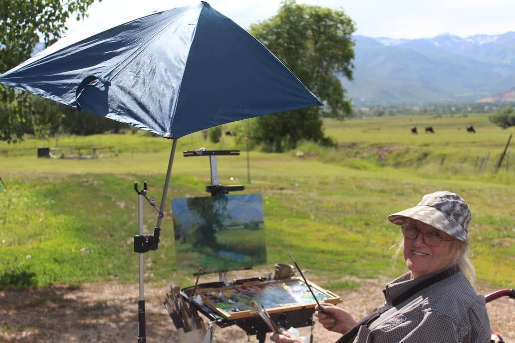 Linda Farrar painting en plein air in a green meadow with mountains in the distance, easel and palette beside her